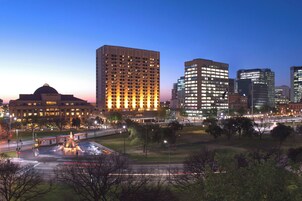 Une vue sur la ville au crépuscule, avec des bâtiments illuminés, une fontaine et un parc.