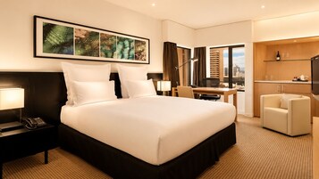 Habitación de hotel con una cama grande, un escritorio, una silla, un televisor, un teléfono, una lámpara, un cuadro en la pared y vistas a la ciudad a través de la ventana.
