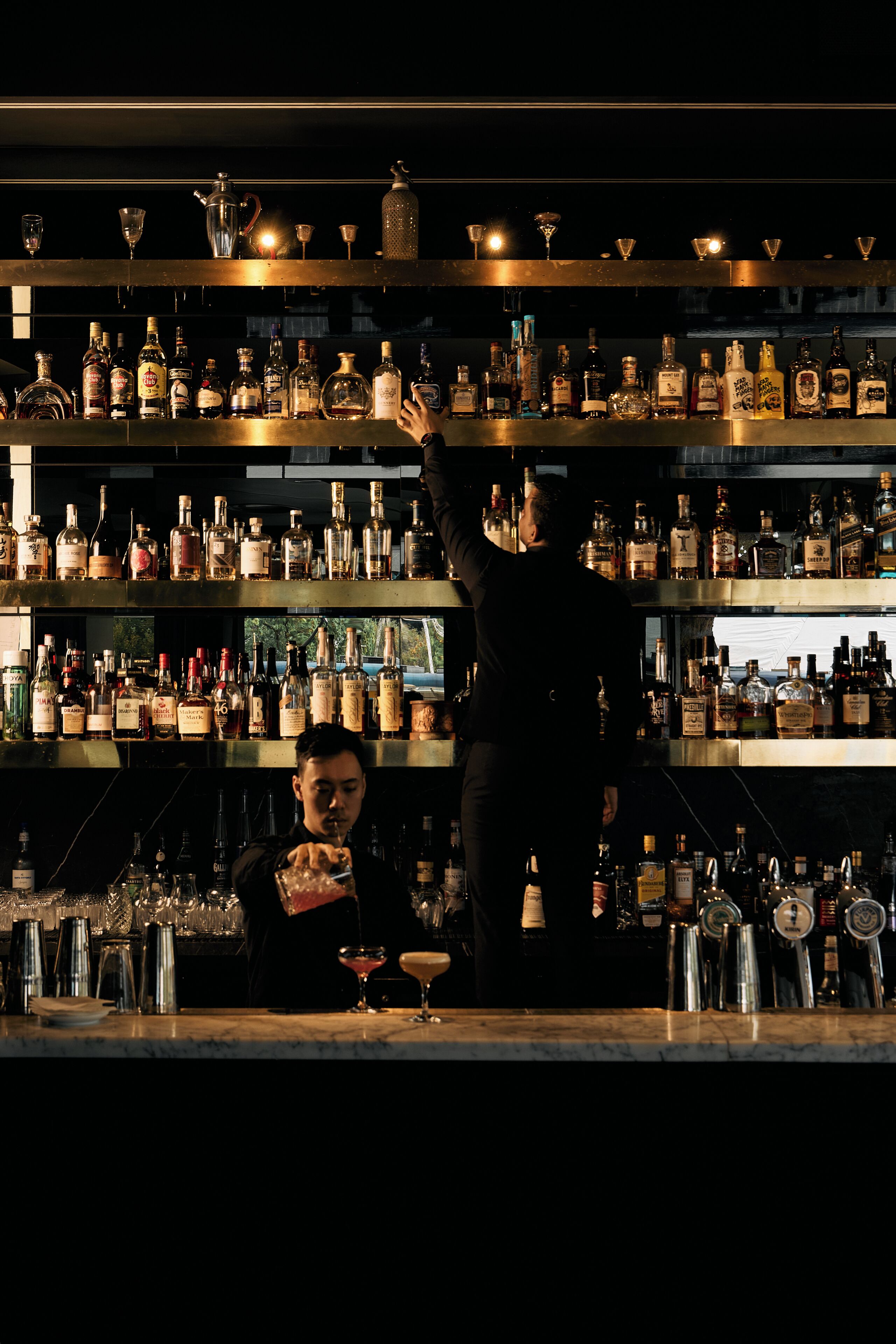 A bar with shelves of liquor bottles and glasses, a bartender preparing a drink, and a customer seated at the bar.