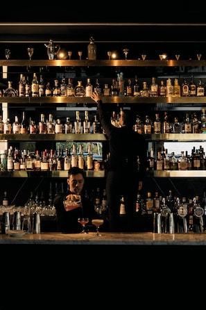 Un bar con estantes llenos de botellas de licor y vasos, un cantinero preparando una bebida, y un cliente sentado en la barra.