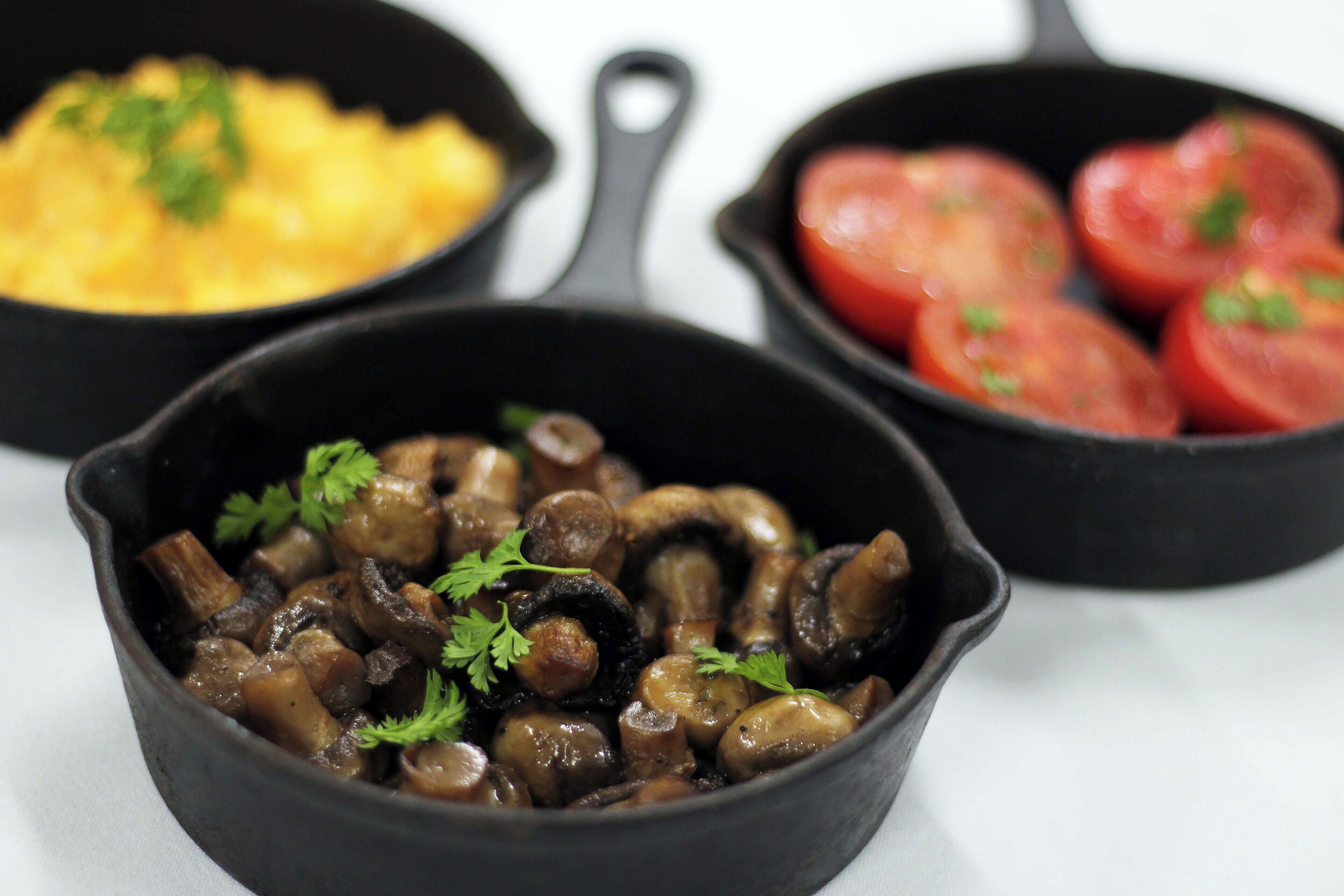 Three black cast iron skillets with food, one containing mushrooms, another with tomatoes, and the third with scrambled eggs.