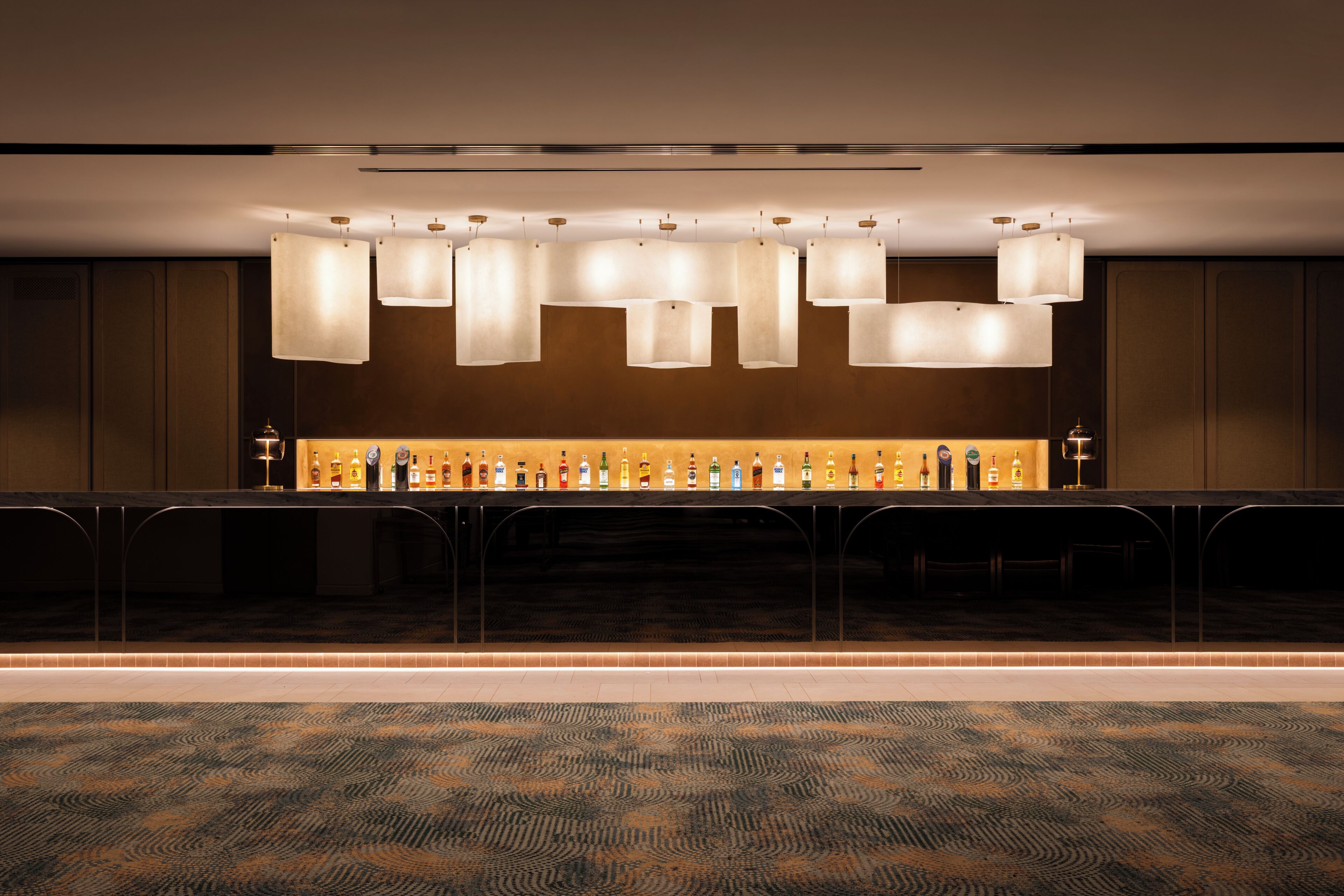 A bar area with a long counter, multiple hanging light fixtures, and a selection of bottles.