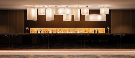 A bar area with a long counter, multiple hanging light fixtures, and a selection of bottles.