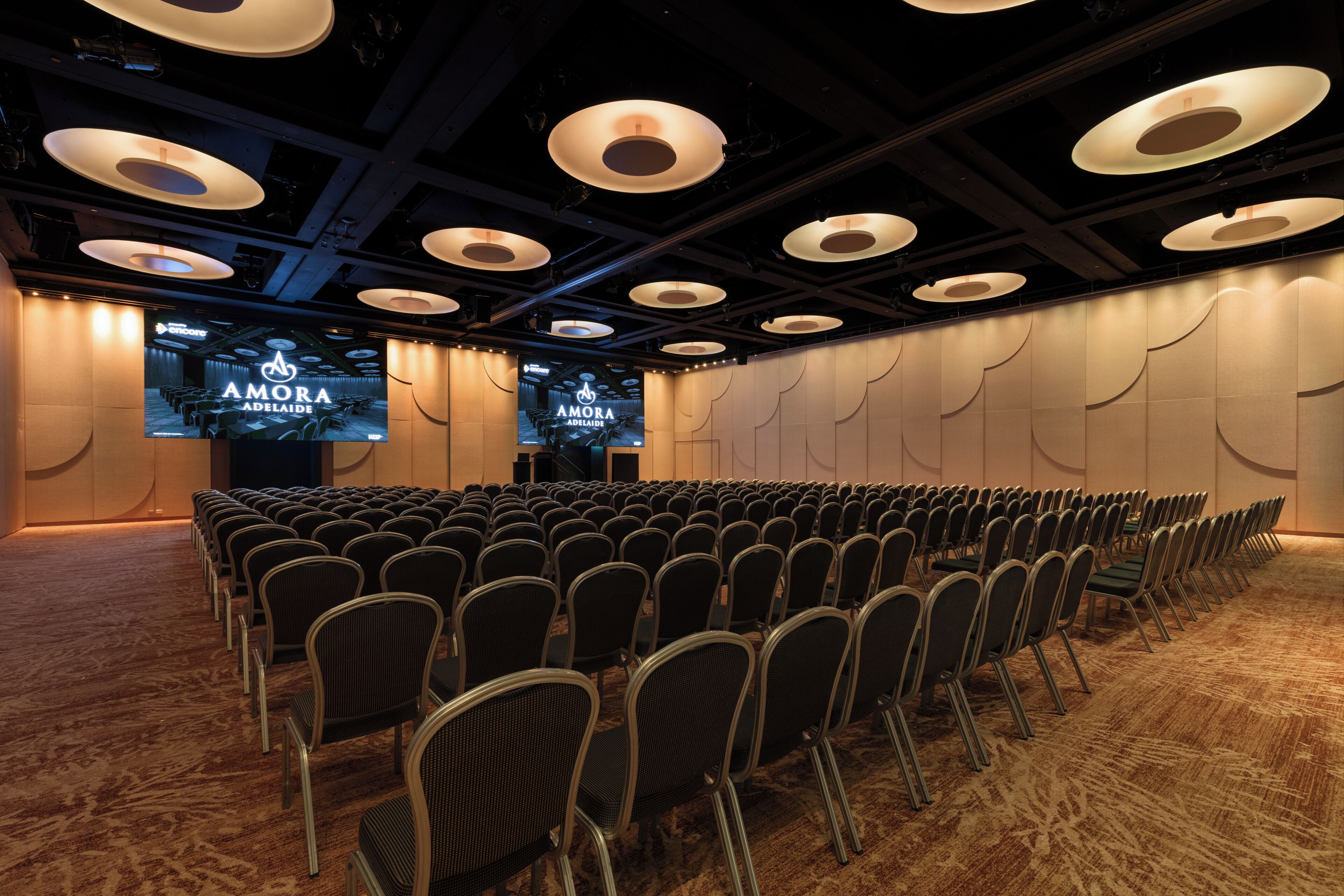 A conference room with rows of chairs, a stage, and a large screen displaying the 'AMORA ADELAIDE' logo.