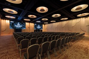 A conference room with rows of chairs, a stage, and a large screen displaying the 'AMORA ADELAIDE' logo. - Amora Hotel Adelaide (Adelaide)