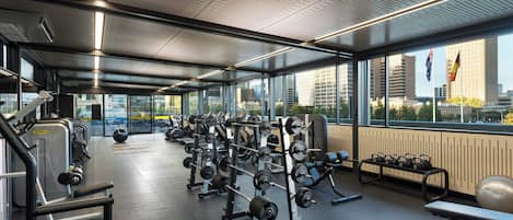 A gym with weightlifting equipment, benches, and a view of the city through large windows.