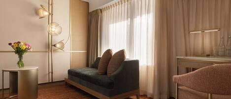Una habitación de hotel moderna con sofá, una mesita de noche con un florero, un escritorio con lámpara y un amplio ventanal con cortinas.