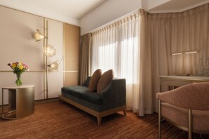 Quarto de hotel moderno com sofá, mesa lateral com um vaso de flores, escrivaninha com luminária e uma grande janela com cortinas.