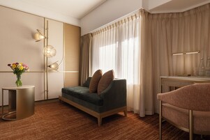 Et moderne hotelværelse med sofa, et sofabord med blomster, et skrivebord med lampe og et stort vindue med gardiner.