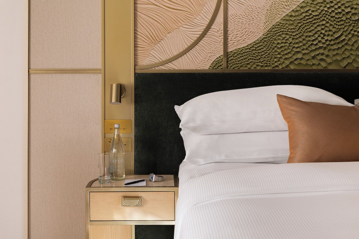 Una habitación de hotel con cama, mesita de noche, botella de agua, vaso y bolígrafo.