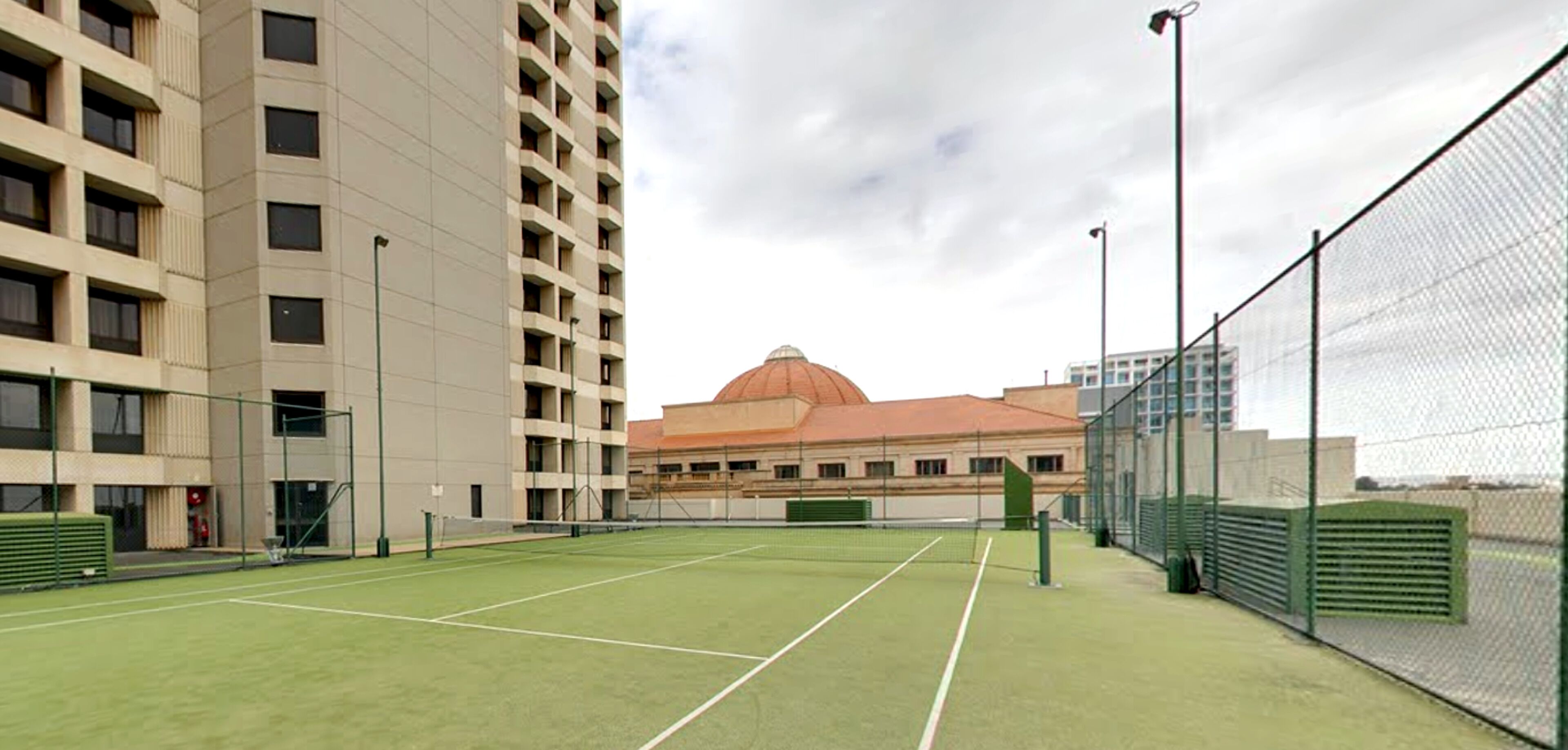 En udendørs tennisbane med net, omgivet af et hegn, og beliggende ved siden af en bygning med et kuppelformet tag.