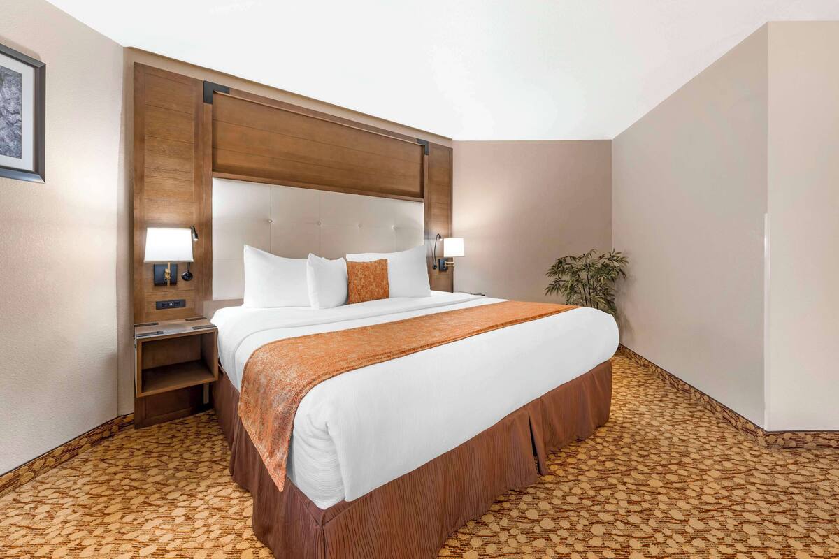 A hotel room with a large bed, wooden headboard, two bedside tables with lamps, a nightstand, and a patterned carpet.