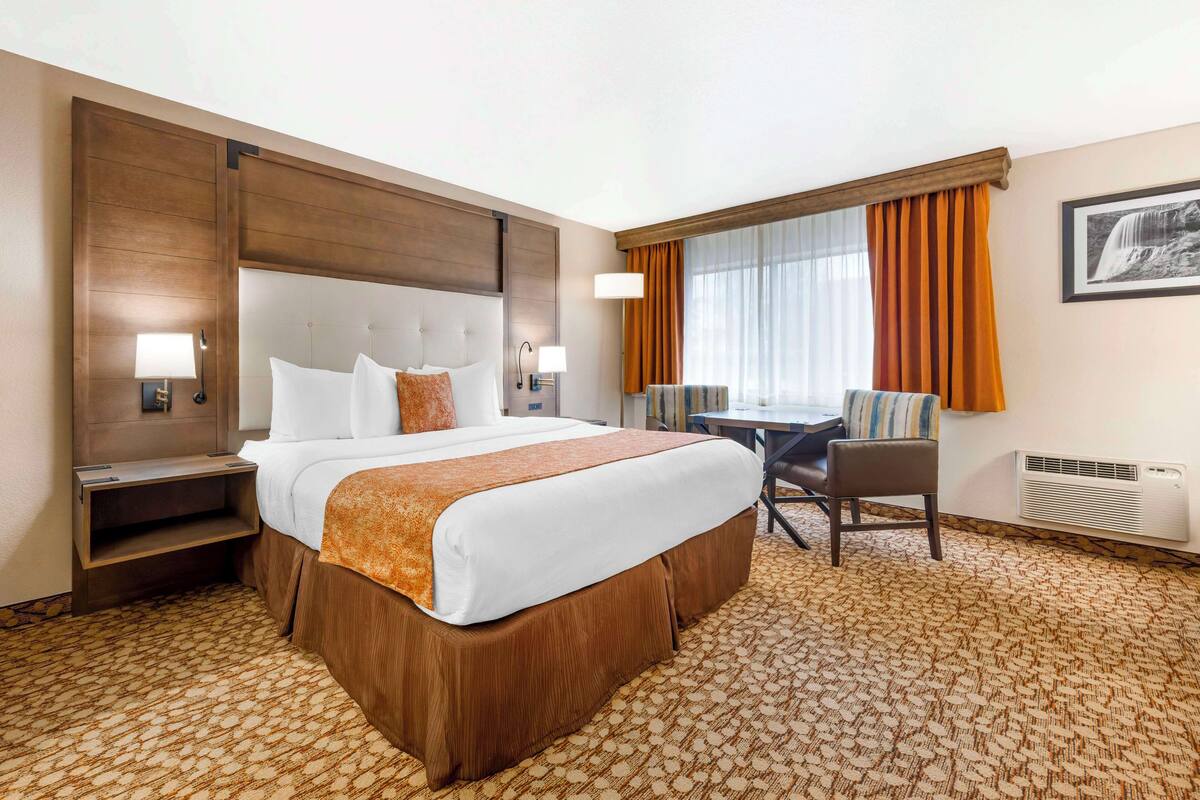 A hotel room with a large bed, a desk, and a chair. There is a window with curtains, a wall-mounted heater, and a framed picture on the wall.