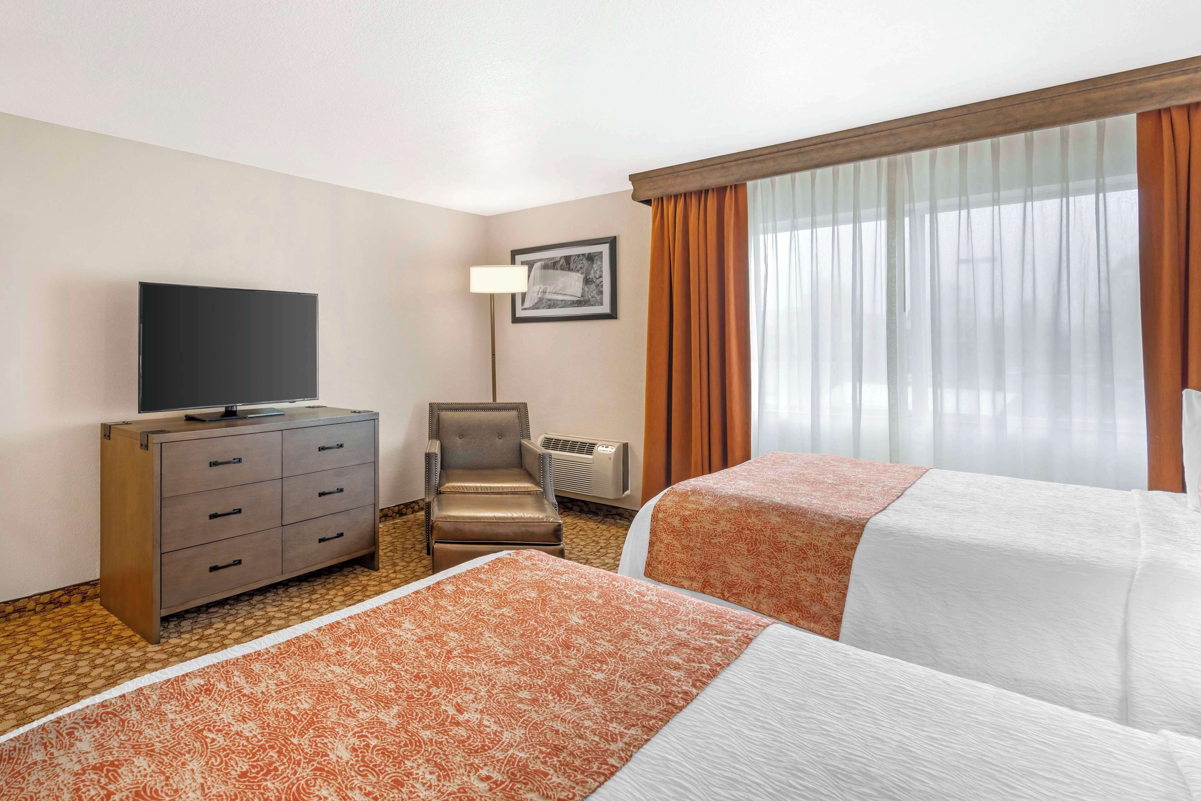 Habitación de hotel con dos camas, un televisor en una cómoda, una silla y un ventanal con cortinas.