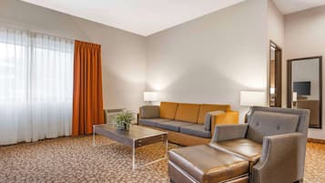 A hotel room with a sofa, armchair, ottoman, coffee table, and a lamp.