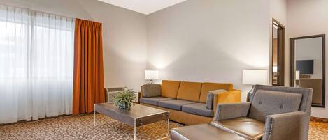 A hotel room with a sofa, armchair, ottoman, coffee table, and a lamp.