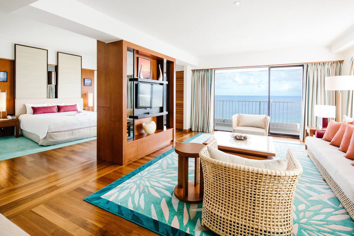 Quarto de hotel com cama, televisão, sofá, poltrona de vime e varanda com vista para o mar.