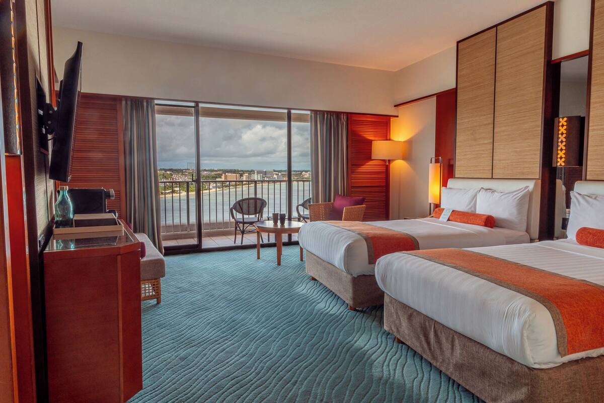 Quarto de hotel com duas camas, uma escrivaninha, uma cadeira e varanda com vista para a cidade.