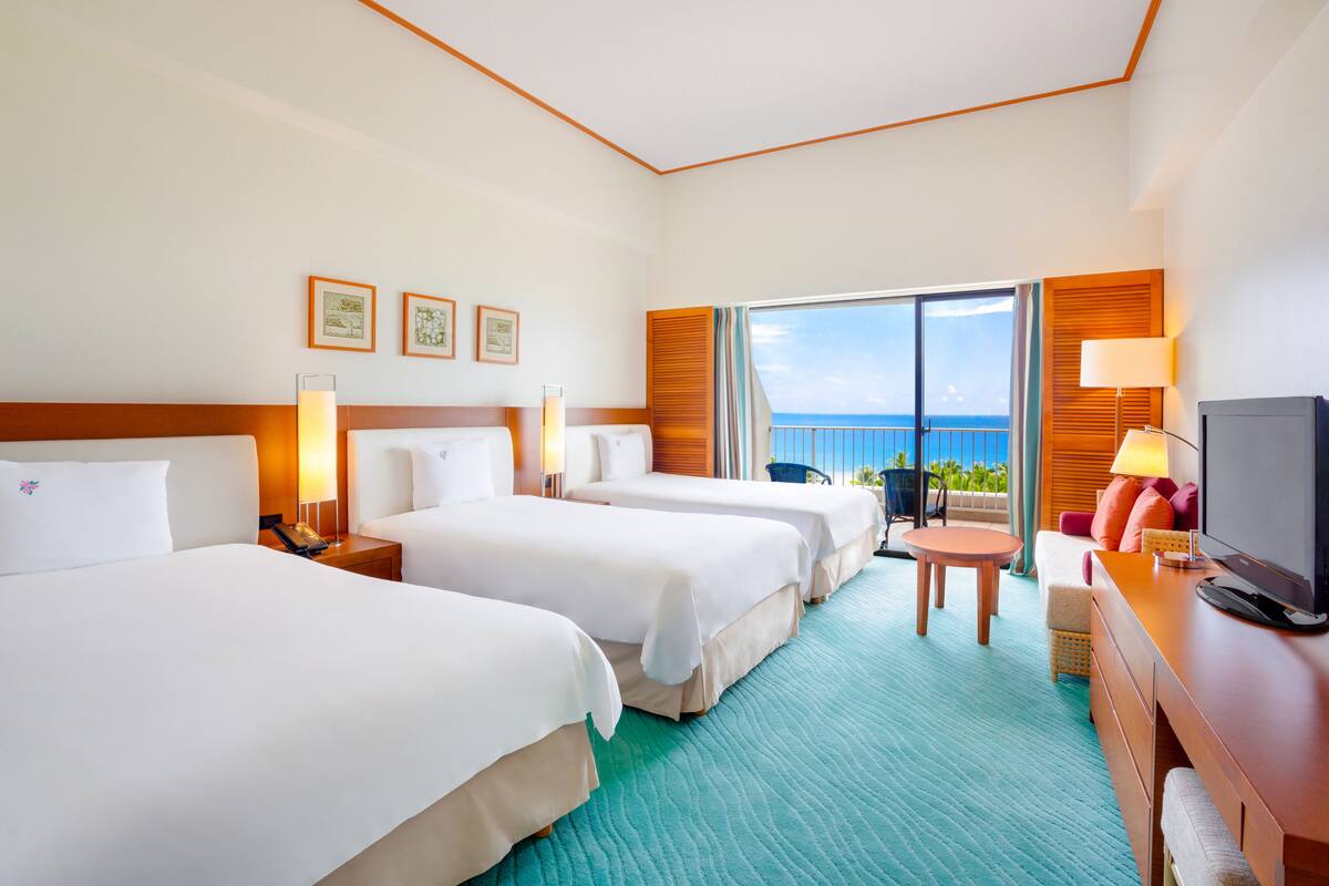 Quarto de hotel com duas camas, uma escrivaninha, uma televisão e varanda com vista para o mar.