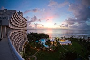 A modern hotel with a curved facade overlooking a pool area and the ocean at sunset.