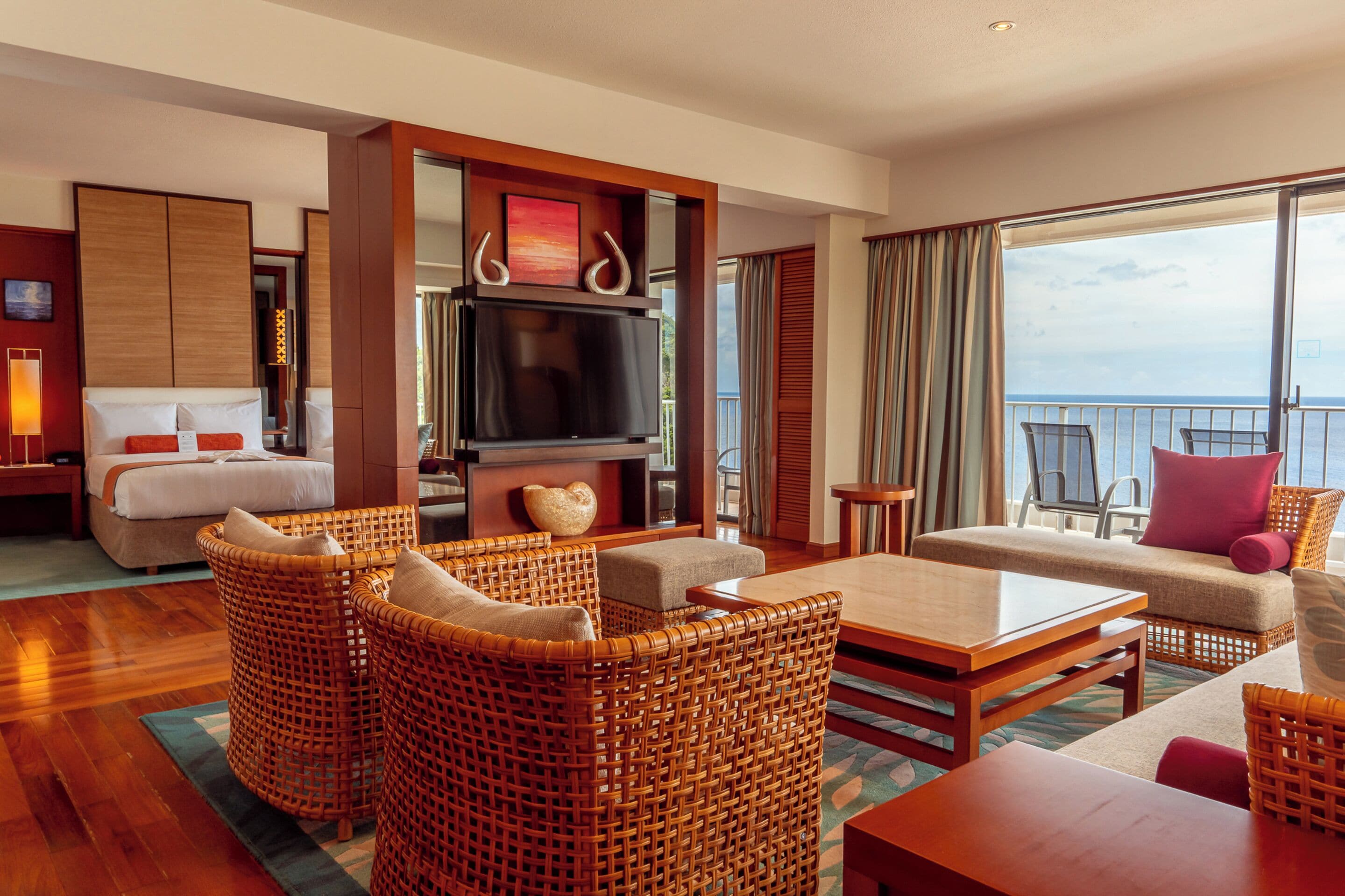 A hotel room with a bed, a TV, wicker furniture, a balcony with a view of the sea, and a large window.