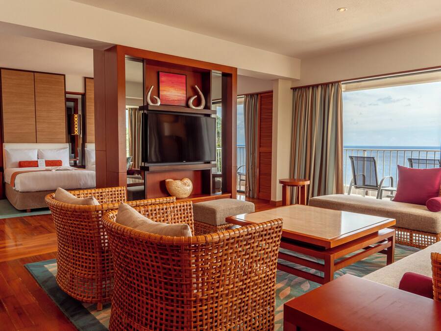 A hotel room with a bed, a TV, wicker furniture, a balcony with a view of the sea, and a large window.
