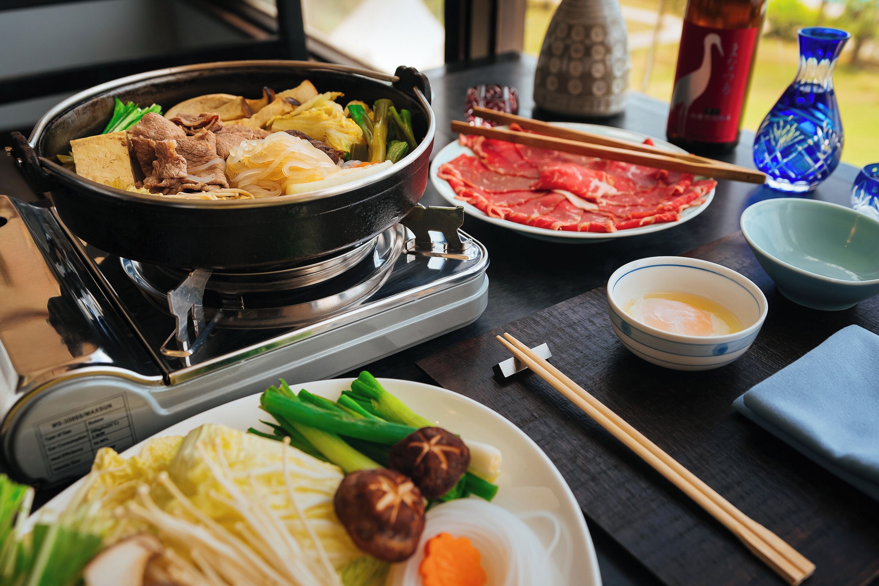 A dining table with a hot pot, raw meat, vegetables, and a bowl of egg.