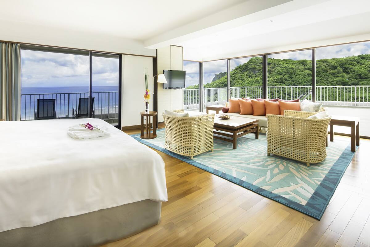 Quarto de hotel com uma cama grande, uma varanda com vista para o mar e uma área de estar com móveis de vime.