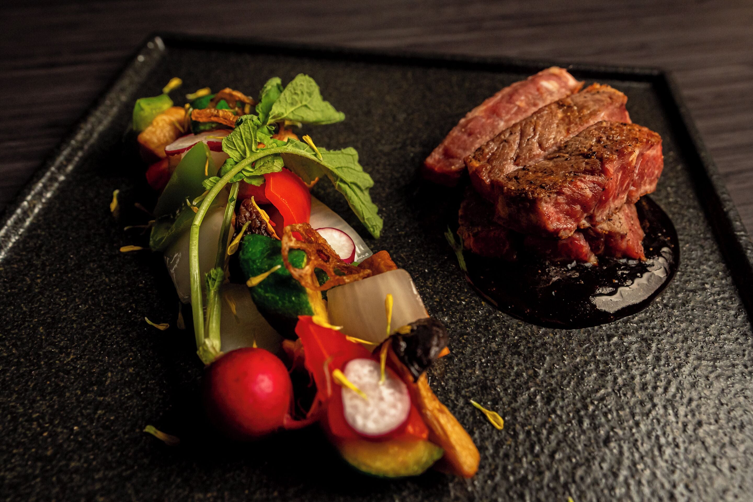 A steak with a dark sauce on a black plate, accompanied by a colorful vegetable dish.