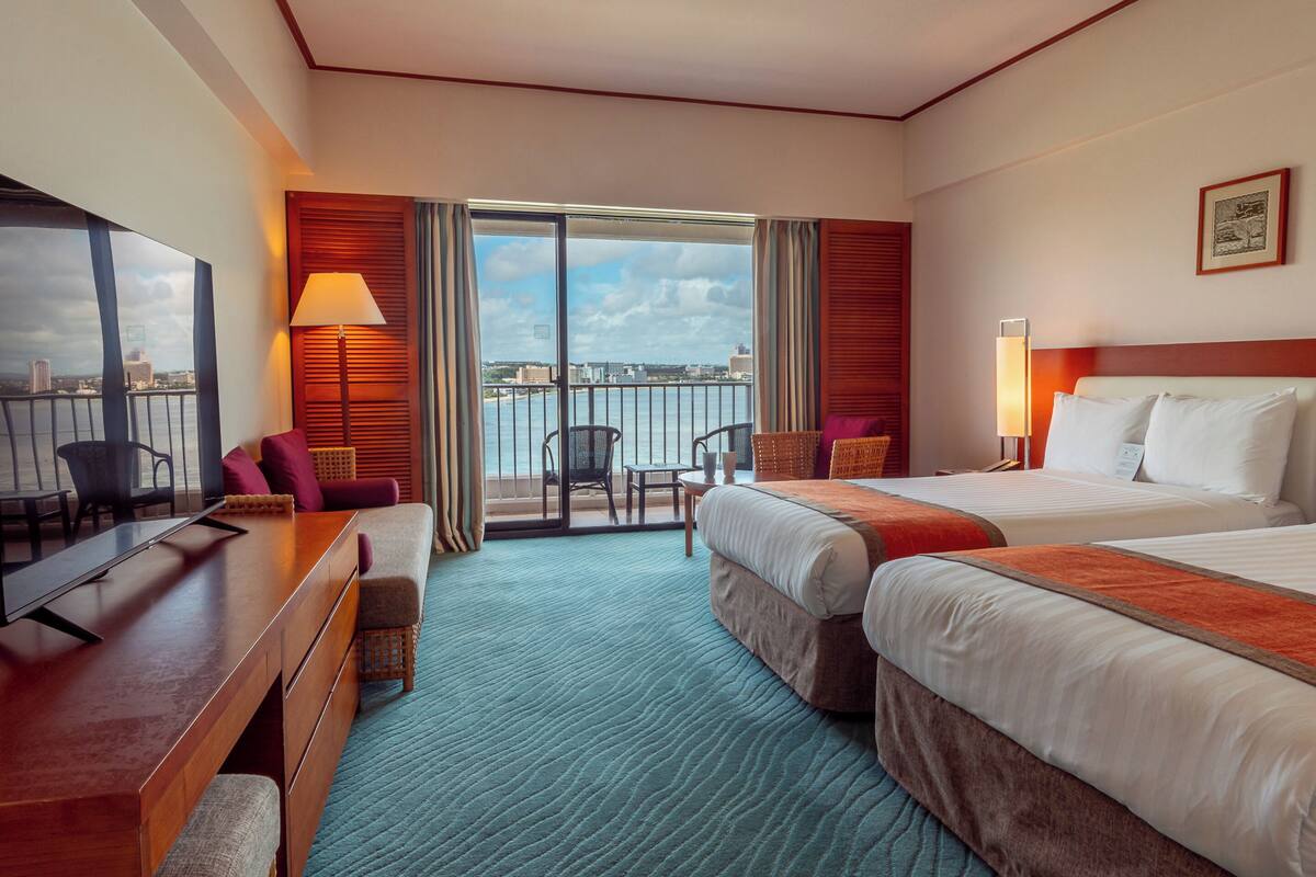 Quarto de hotel com duas camas, uma escrivaninha, televisão e varanda com vista para a cidade.
