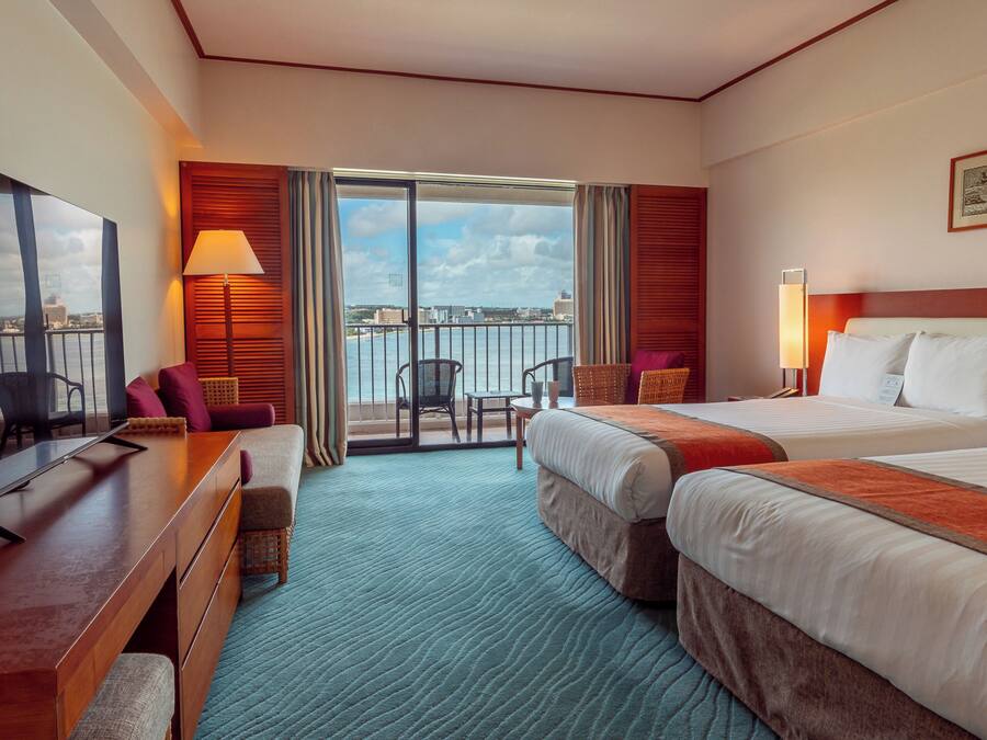 Een hotelkamer met twee bedden, een bureau, een televisie en een balkon met uitzicht op de stad.
