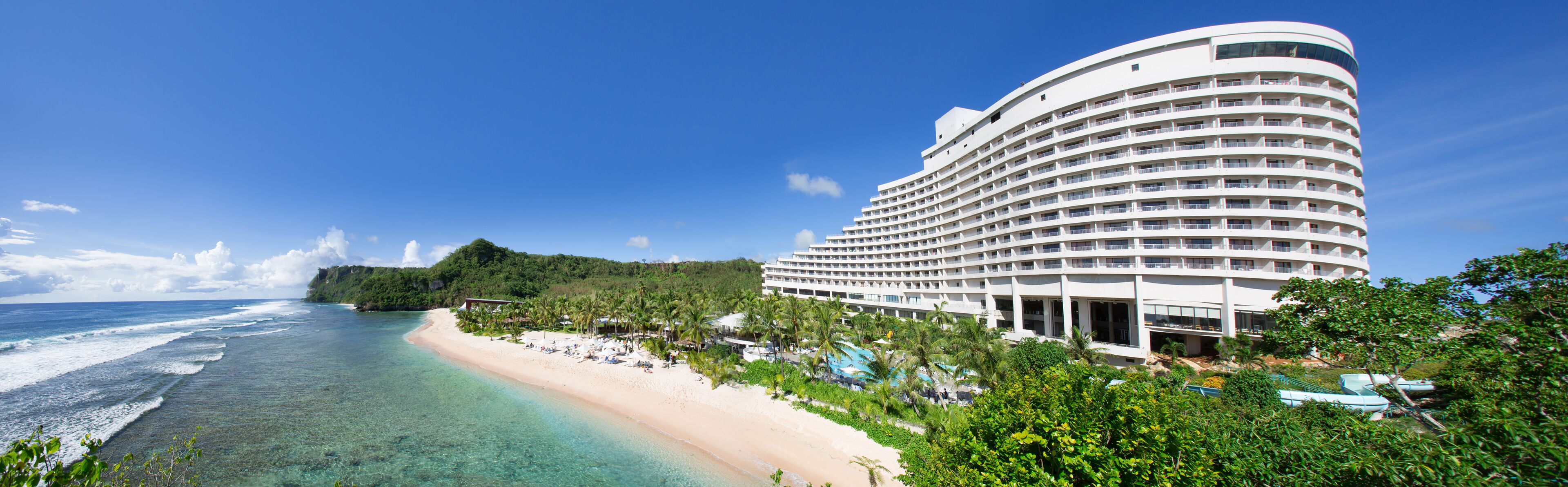 Un hotel frente a la playa con una estructura curva, una piscina y una playa con palmeras.