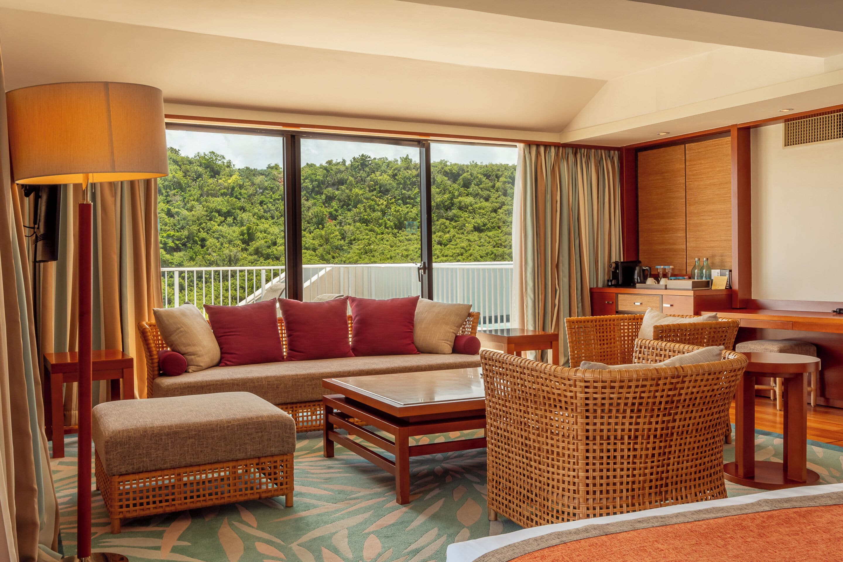 A hotel room with a sofa, ottoman, wicker chairs, a coffee table, and a view of a lush green landscape through large windows.