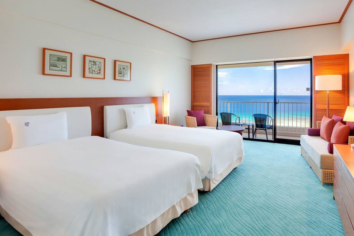 Quarto de hotel com duas camas, varanda com vista para o oceano e área de estar com mesa e cadeiras.