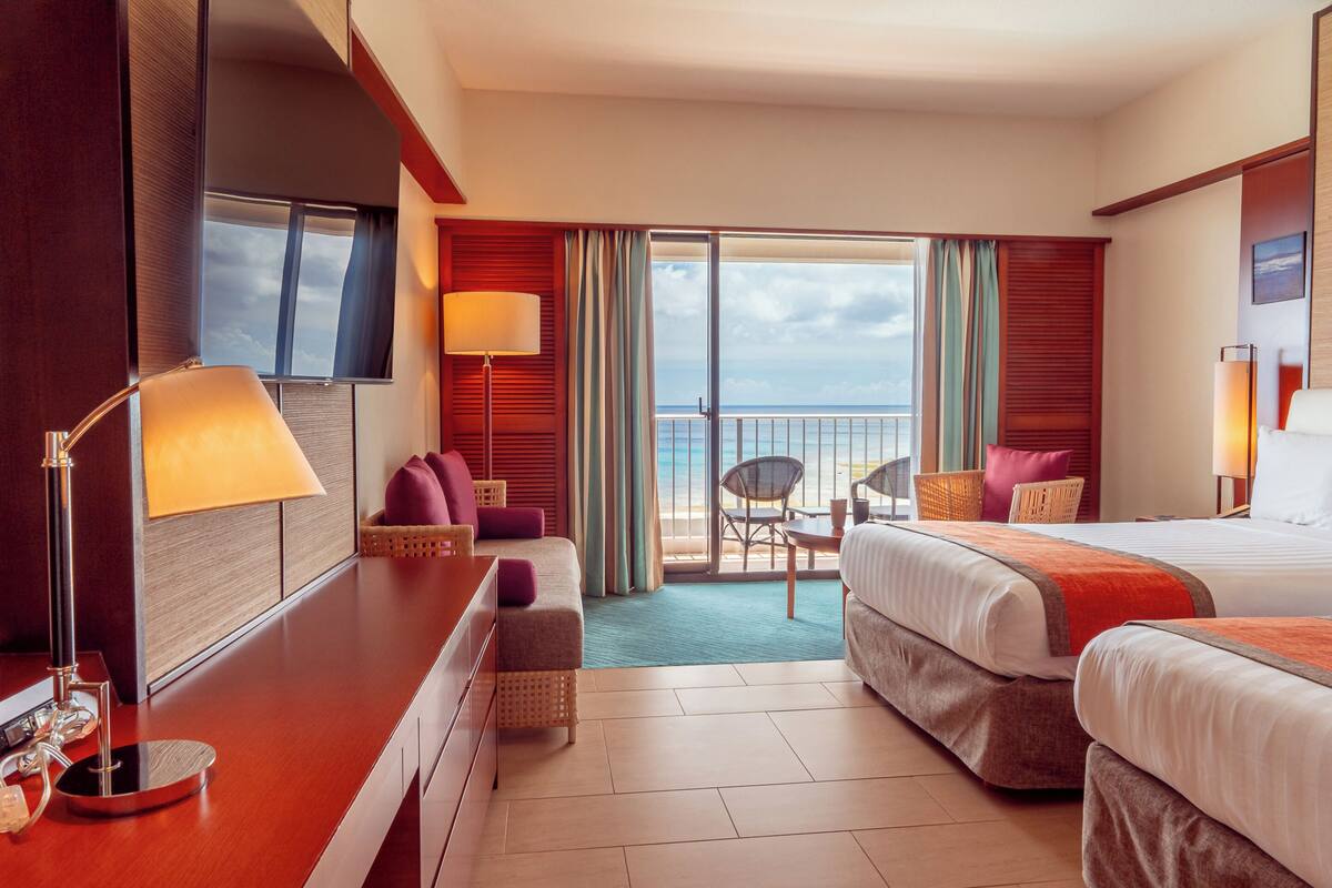 Quarto de hotel com duas camas, uma escrivaninha, uma cadeira, varanda com vista e televisão.