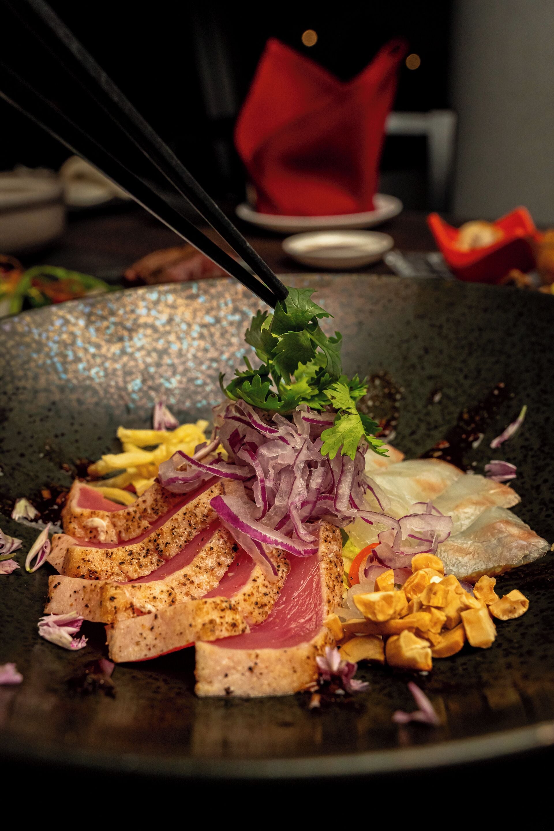 A close-up of a dish with sliced meat, vegetables, and herbs on a dark plate.