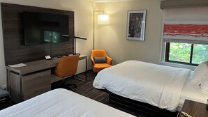 A hotel room with two beds, a desk with a chair, a TV, a lamp, and a window with a view of greenery. - Rodeway Inn Little Falls (Little Falls)