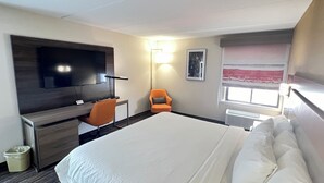 A hotel room with a large bed, a desk with a chair, a flat-screen TV mounted on the wall, and a window with a view.