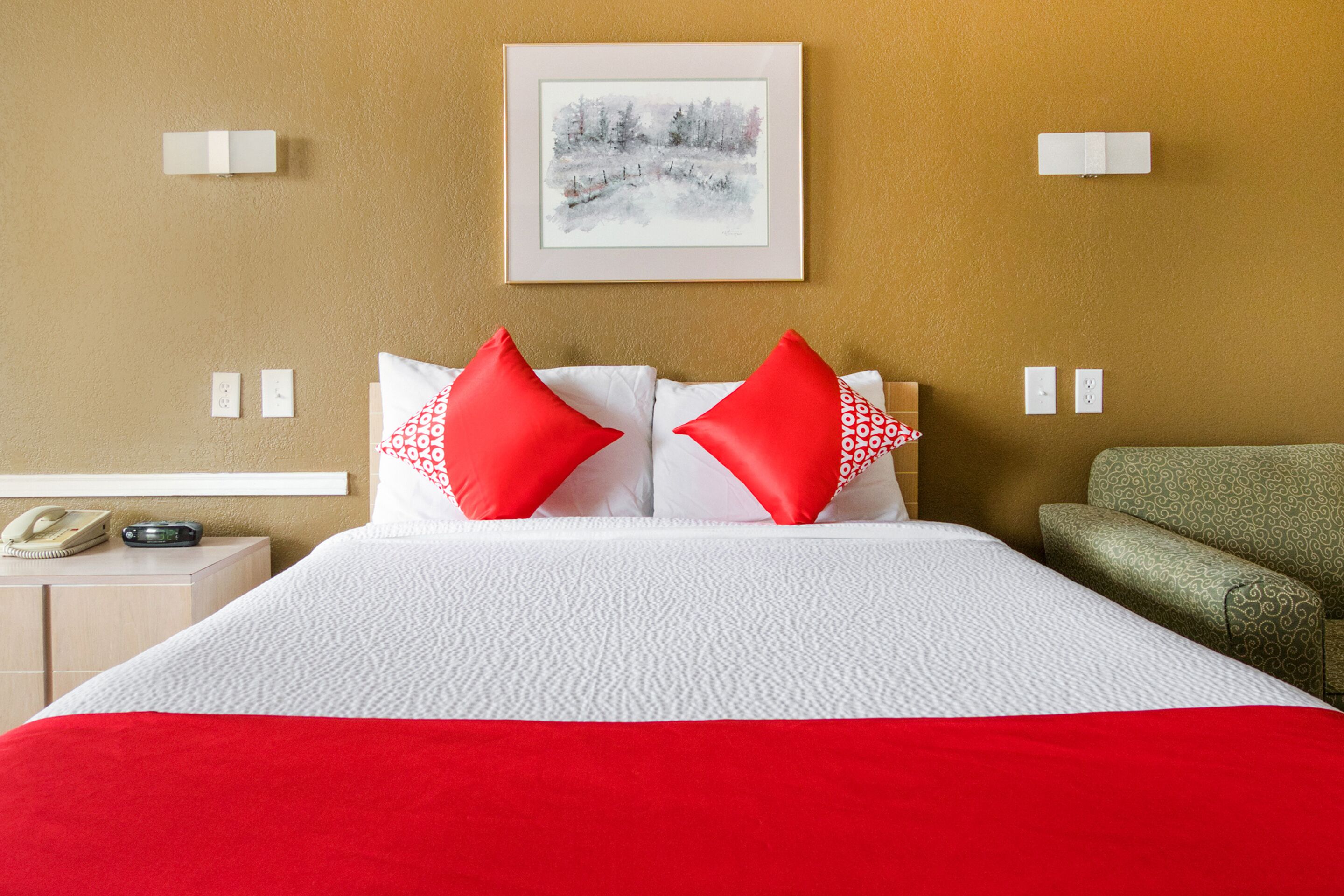 Een hotelkamer met een bed, twee rode kussens, een ingelijst schilderij aan de muur, nachtlampjes, een telefoon en een klok.