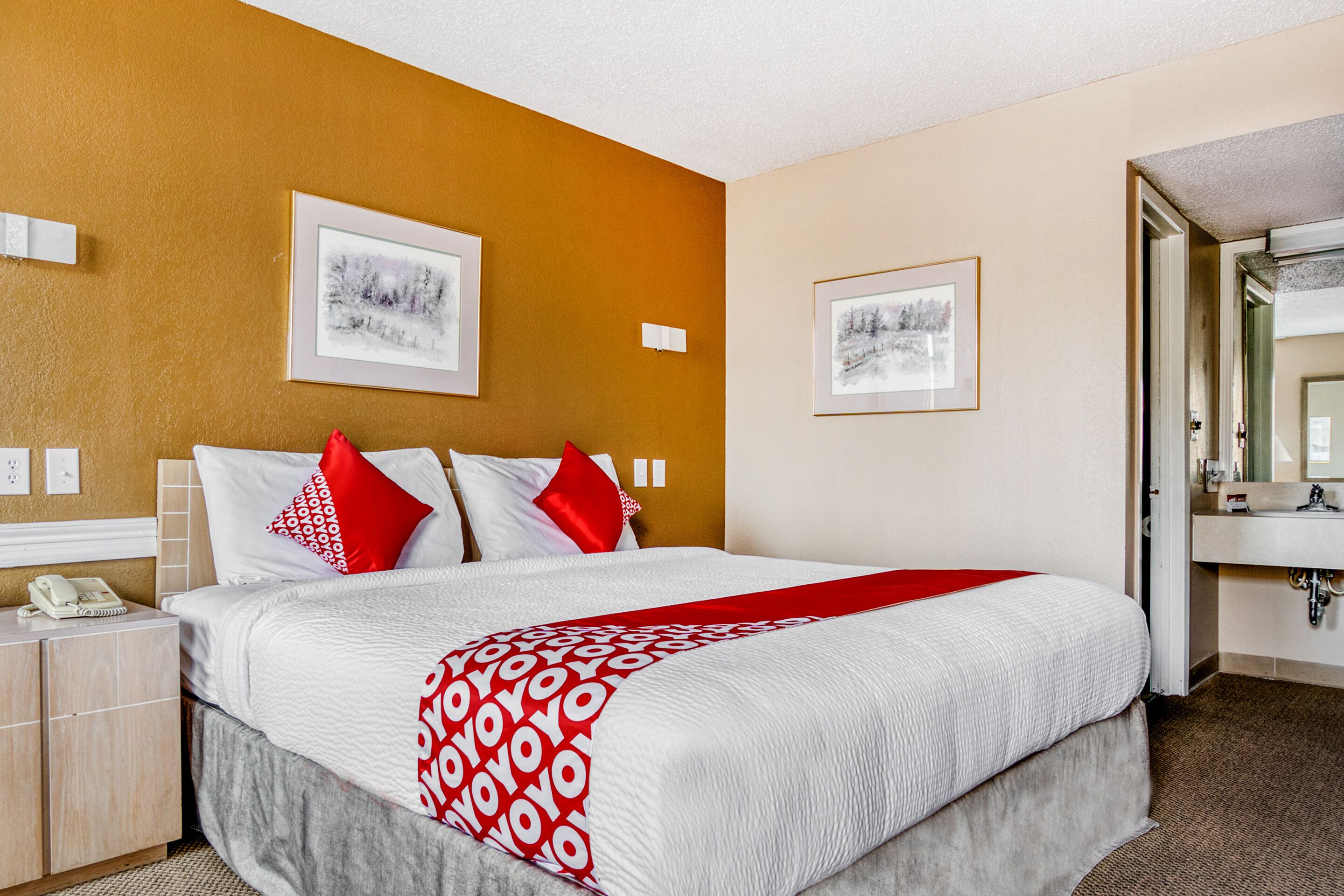 A hotel room with a large bed, two red pillows, a white bedspread, and two framed pictures on the wall.