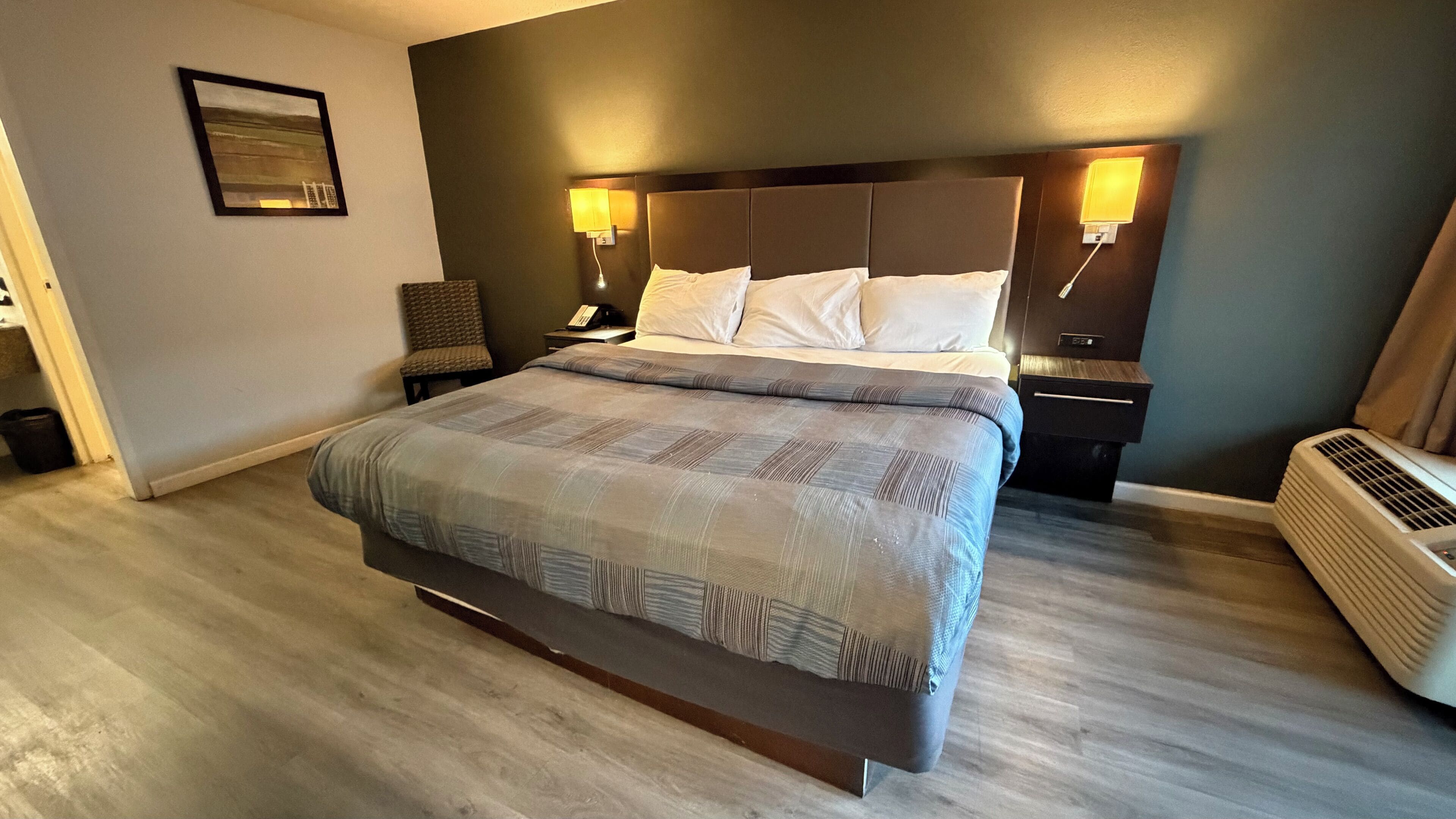 A modern hotel room with a large bed, a chair, a nightstand, and a wall-mounted heater.