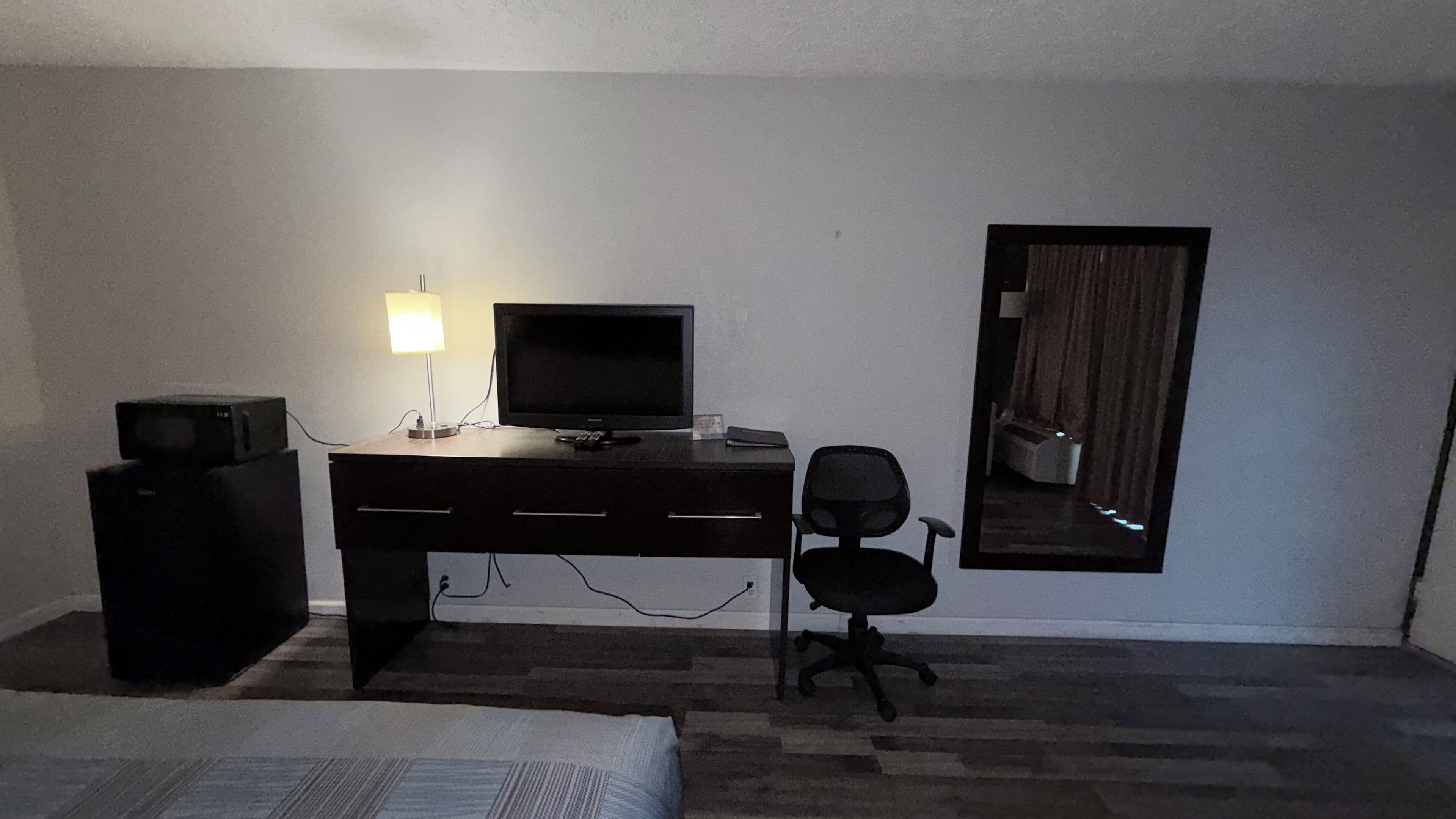 Ein kleiner, schwach beleuchteter Raum mit einem Bett, einem Schreibtisch mit Computer und Lampe sowie einem Stuhl.