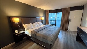 Ein Hotelzimmer mit einem großen Bett, Nachttischlampen, einem Nachttisch, einem Schminktisch und Blick auf Gebäude durch das Fenster.