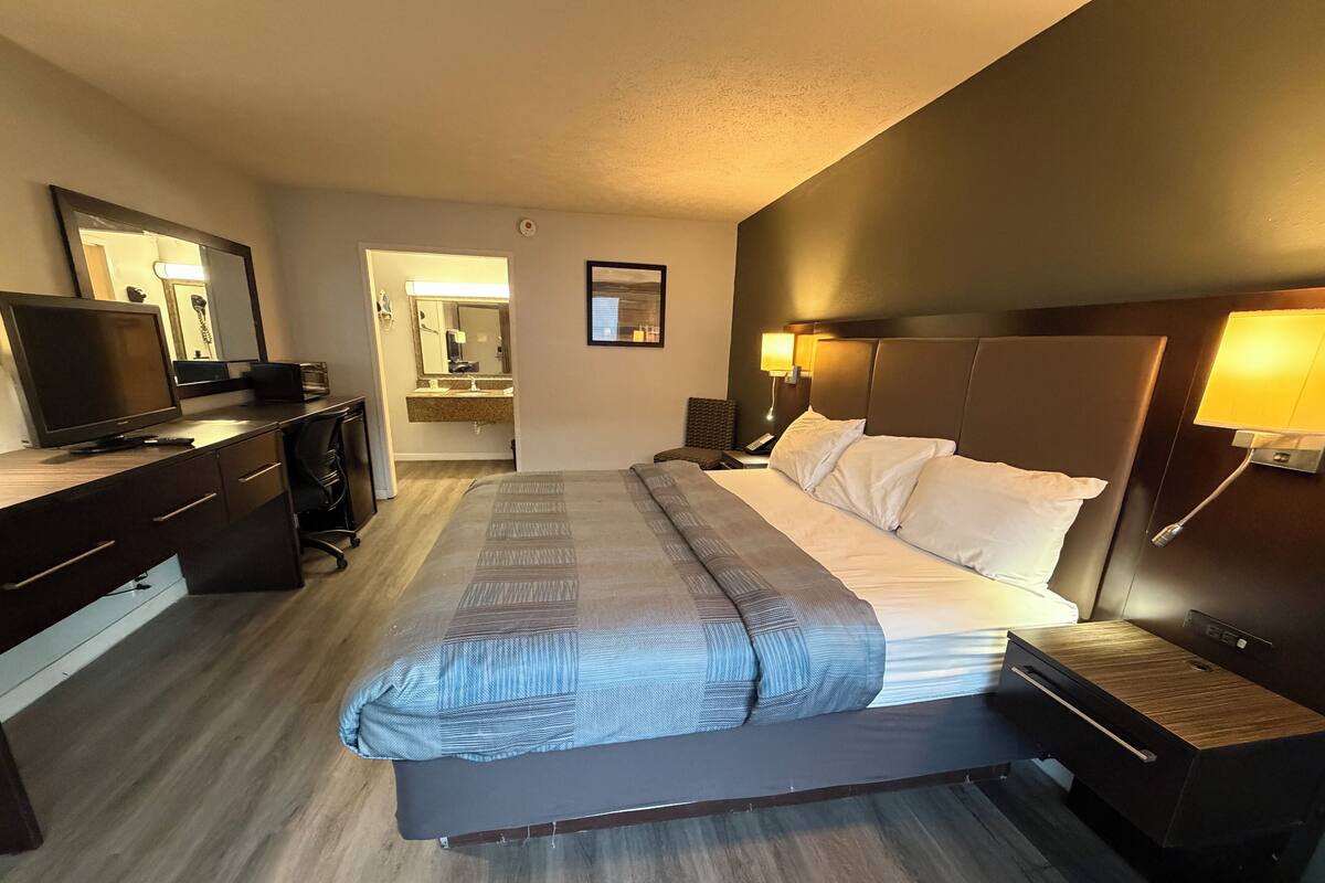 A hotel room with a large bed, a desk with a computer, a TV, and a bathroom visible through an open door.