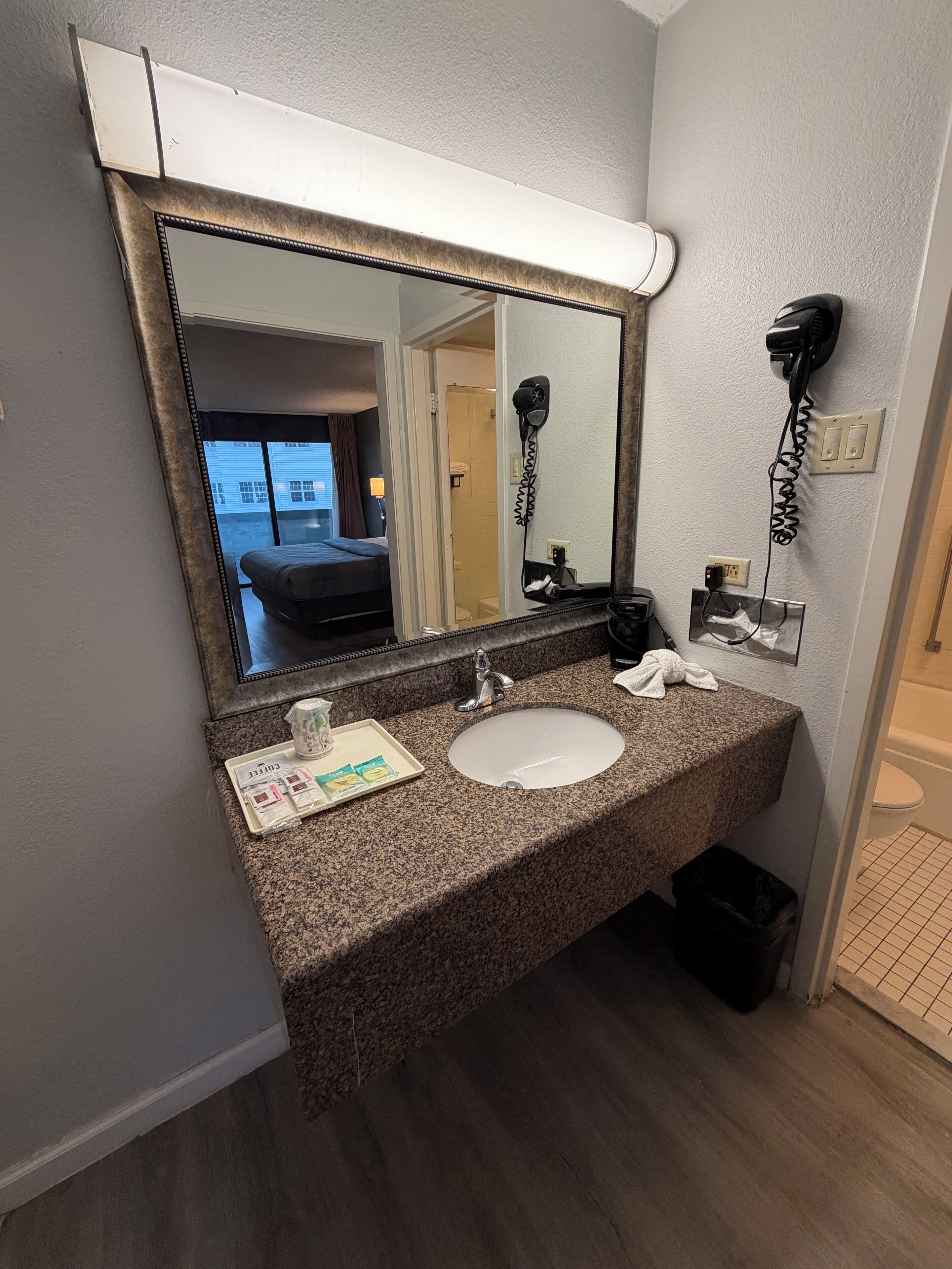 Une salle de bain d’hôtel avec un lavabo doté d’un plan de travail en granit, un grand miroir, un sèche-cheveux et un porte-serviettes.