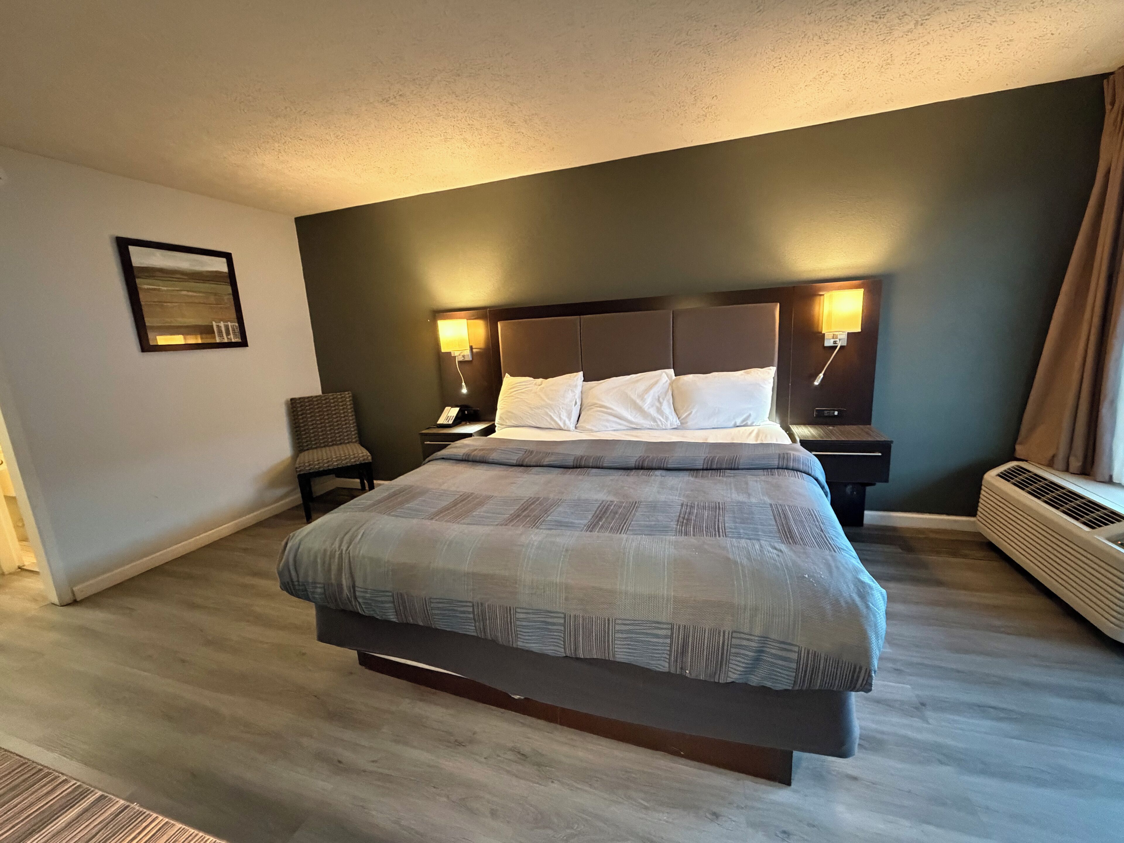 A hotel room with a large bed, two bedside tables, a chair, a wall-mounted lamp, and an air conditioning unit.