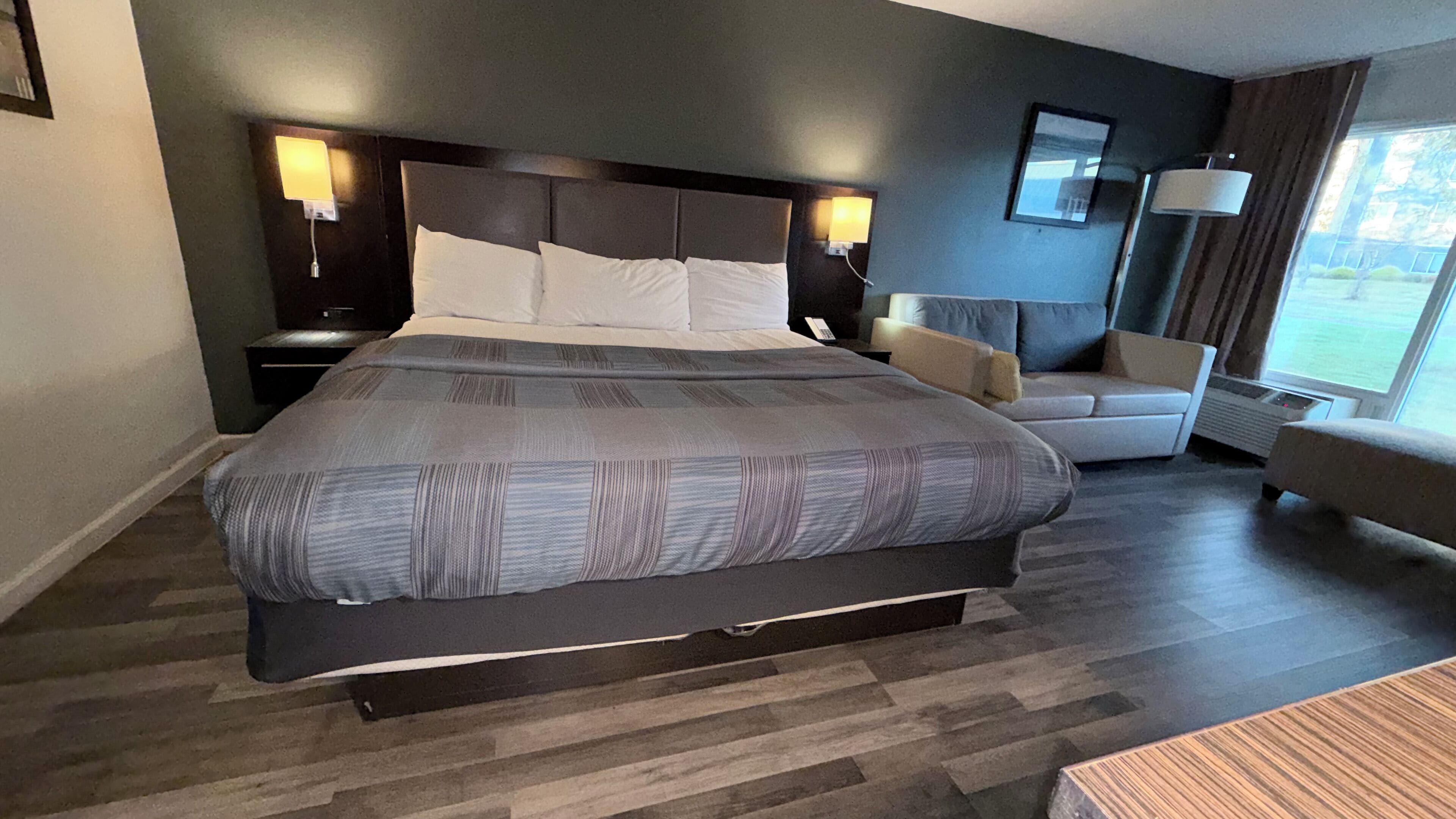 A large bed with a headboard, two bedside tables with lamps, a chair, and a large window with curtains.
