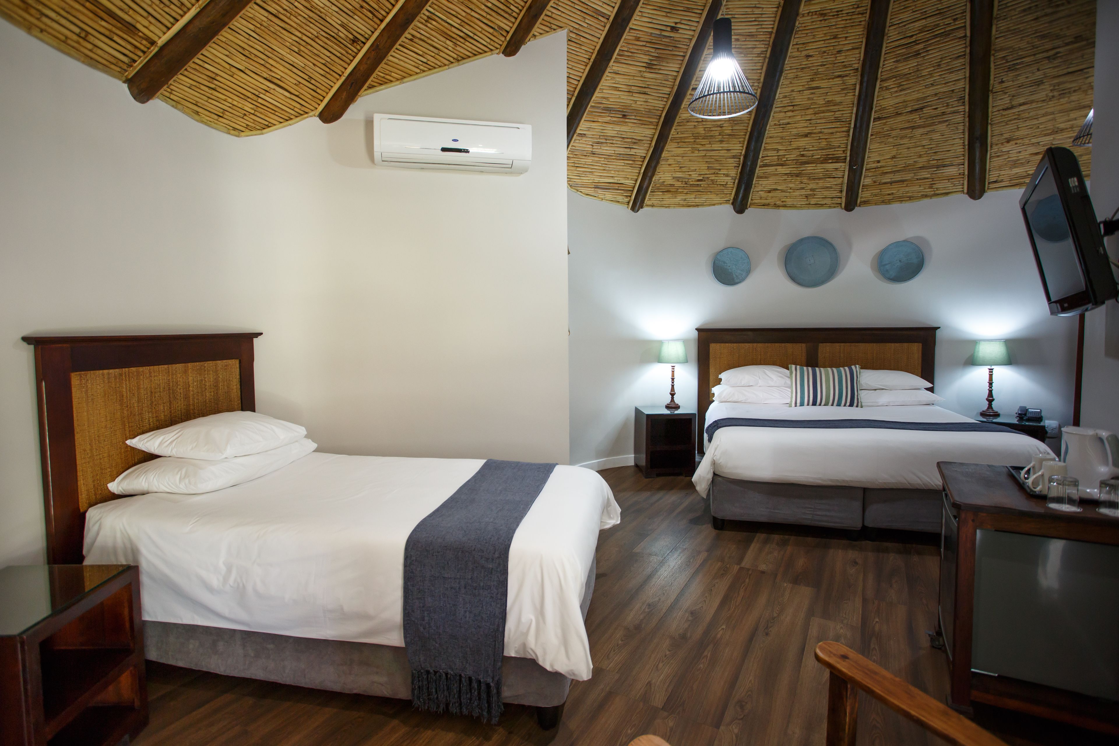 Habitación de hotel con dos camas, piso de madera, ventilador de techo, televisor y una mesita con bandeja.