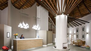 Un área de recepción con un mostrador de madera, lámparas colgantes, un pilar blanco grande y un sofá con una butaca de mimbre.