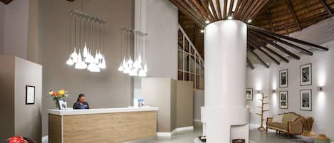 A reception area with a wooden reception desk, hanging pendant lights, a large white pillar, and a sofa with a woven chair.